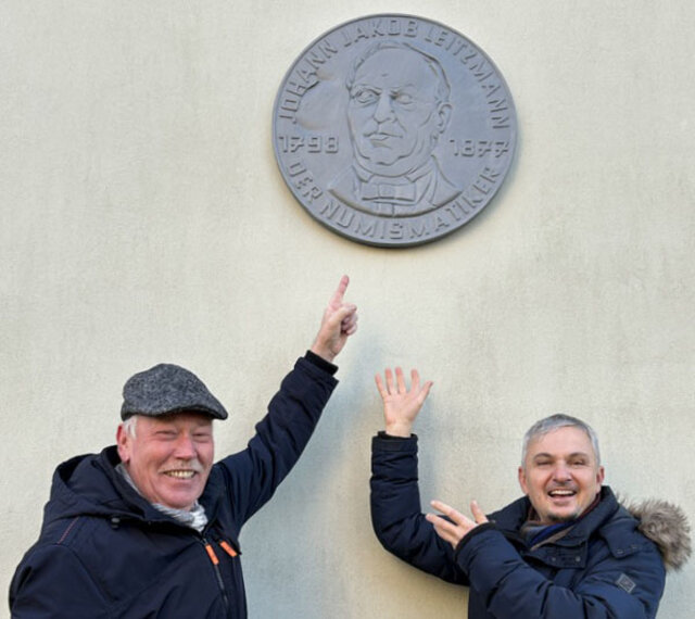 Ortsteilbürgermeister Siegfried Schmidt (l.) und Dr. Hans-Dieter Dörfler, Leiter Museum und Archiv, unter der restaurierten Leitzmann-Plakette. Zwei Männer stehen mit lachenende Gesichern vor einer hellen Hausfassade und zeigen nach oben auf eine große runde Plakette, die an der Hauswand angebracht ist.