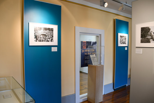 Raum der Sonderausstellung im Museum angeleuchteten Fotos an den Wänden sowie beleuchteten Vitrinen mit Ausstellungsstücken.