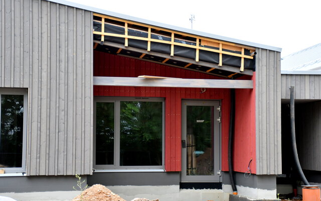 Ansicht des Neubaus der Kita Flax & Krümel in Sömmerda. Zu sehen ist die moderne Fassade aus grauen und roten Holzpaneelen mit großen Fenstern und einem Eingangsbereich.