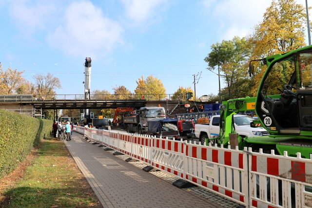 Die jetzige Baustellensituation von der Stadtmitte kommend: Der Fußweg im Baustellenbreich ist mit rot-weißen Baaken von der Straße abgetrennt. Hinter den Baaken stehen verschiedene Baufahrzeuge auf der Straße. Links neben dem Fußweg befindet sich eine Grünfläche. Eine kleine Gruppe Fußgänger steht in einer Entfernung der Brücke und schaut den Arbeitenden zu. Hinter der Brücke ragt ein großer Kran auf.
