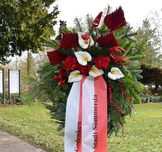 Auf einem Ständer steht ein aus verschiedenem Tannengrün gewundener Kranz. In den Kranz sind Blumen in Rot und Weiß - den Stadtfarben von Sömmerda - gesteckt. Dazu wurden noch einige rote Deko-Elemente auf dem Kranz platziert. Außerdem ziert ihn eine reot-weiß gestreifte Schleife. Im Bild sind auf den langen Schleifenbändern die Worte Gedenken sowie Stadt Sömmerda zu lesen.