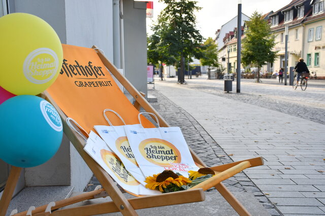 Heimat shoppen in der Sömmerdaer Innenstadt Im Vordergrund stehen zwei orangefarbene Liegestühle mit mehreren „Heimat shoppen“-Einkaufstüten und Sonnenblumen. Daneben sind gelbe, pinkfarbene und türkisfarbene Luftballons befestigt. Im Hintergrund ist die Fußgängerzone der Sömmerdaer Innenstadt mit Geschäften, Bäumen, Sitzgelegenheiten und einer Radfahrerin zu sehen.