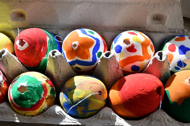 Bunte, handbemalte Ostereier liegen nebeneinander in einem Eierkarton. Die Eier sind mit verschiedenen Farben und Mustern verziert, darunter Punkte, Streifen und Blumenmotive. Einige Eier haben Fäden zum Aufhängen. Das Sonnenlicht fällt von der Seite auf die Eier und hebt die leuchtenden Farben hervor.