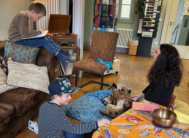 In einem gemütlich eingerichteten Raum mit warmem Licht sitzen zwei Jungen und eine Frau gemeinsam mit einem Hund. Der Junge auf dem braunen Sofa liest konzentriert ein Buch, während der andere Junge auf dem Teppich sitzt und den auf dem Rücken liegenden Hund sanft streichelt. Die Frau sitzt ebenfalls auf dem Boden und schaut lächelnd zu. Umgeben sind sie von bunten Kissen, Decken, einem Plattenspieler, Büchern und einer tibetischen Klangschale auf einem Tisch mit farbenfroher Decke. Die Atmosphäre wirkt ruhig, liebevoll und geborgen.