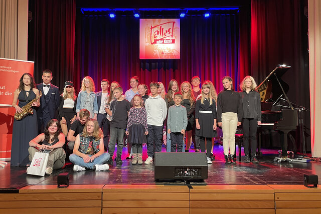 Teilnehmende des Musikwettbewerbs „Alles außer Klassik“ auf der Bühne des Volkshauses Sömmerda Gruppenfoto der teilnehmenden Kinder und Jugendlichen beim Musikwettbewerb „Alles außer Klassik“ im Volkshaus Sömmerda. Auf der Bühne stehen und sitzen junge Musikerinnen und Musiker verschiedener Altersgruppen vor einem roten Bühnenvorhang, im Hintergrund ist das Logo „Alles außer Klassik“ zu sehen. Ein Flügel und eine Saxophonspielerin sind ebenfalls im Bild.