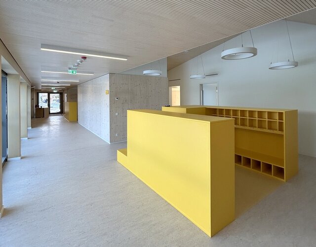 Ein heller und weitläufiger Flurbereich der neuen Kita. Links ist er von bodentiefen Fenstern begrenzt. Die Wände auf der rechten Seite sind mit hellem Holz vertäfelt. Im Vordergrund steht eine knallgelbe Garderobenkombination aus Holz für die Kindersachen.