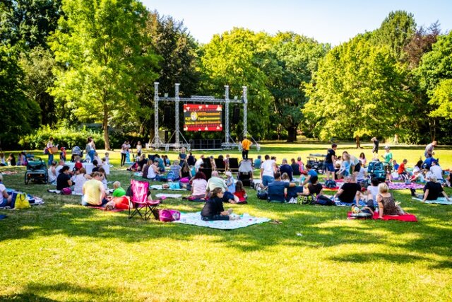 Zahlreiche Familien sitzen bei sonnigem Wetter auf Picknickdecken und Campingstühlen auf einer großen Wiese im Stadtpark Sömmerda. Im Hintergrund steht eine große Leinwand mit dem Schriftzug „Willkommen – KJP-Familiensommerkino – Kino im Stadtpark Sömmerda“. Die Veranstaltung findet unter freiem Himmel bei grünem Baumbestand statt.