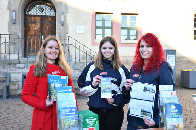 Drei Frauen stehen nebeneinander an einem Infostand auf dem Sömmerdaer Fahrradfrühling vor dem Rathaus. Sie halten verschiedene Flyer und Karten zu Radwegen in der Region Thüringer Becken in den Händen und lächeln in die Kamera. Auf dem Tisch vor ihnen liegen Prospekte zu Radtouren, z. B. zum Gera-, Saale- oder Unstrutradweg. Im Hintergrund ist eine historische Eingangstür mit Steintreppe und Fensterfront zu sehen.