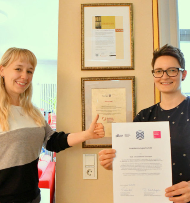 Zwei Frauen stehen vor einem Pfeiler in der Bibliothek, an dem Urkunden hängen. Eine von ihnen hält mit beiden Händen eine weitere Urkunde.