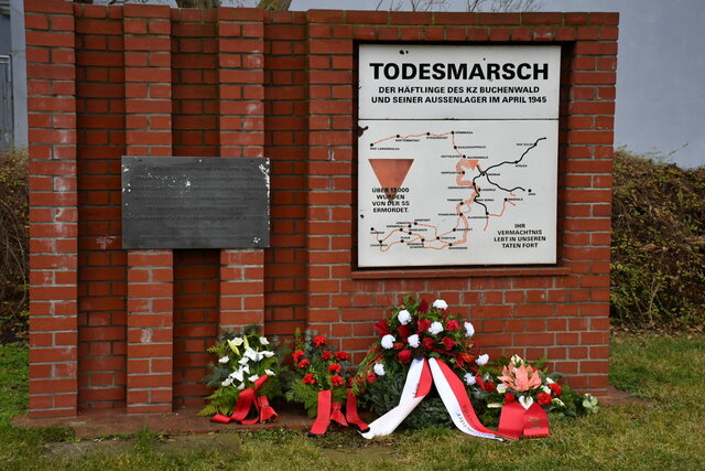 Gedenkort zum Todesmarsch der Häftlinge des KZ Buchenwald und seiner Außenlager im April 1945 auf einer weißen Tafel angebracht auf einer Backsteinsäule. Davor liegen Blume und Kränze zum Gedenken