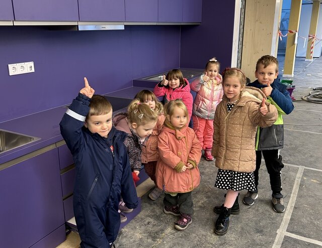 Eine Kindergruppe steht im Neu ihrer Kita vor einer dunkellilafarbenen Kinderküche und winkt in Richtung Fotografin. Im Hintergrund des Fotos sind in regelmäßigen Abständen helle Holzpfeiler zu sehen. Zwischen ihnen wurde rot-weißes Band zur Absperrung gezogen. Der Fußboden ist zum Schutz vor Schmutz noch mit dünnem Filz ausgelegt.