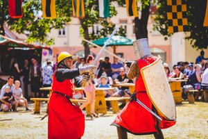 Zwei Darsteller in mittelalterlichen Kostümen und Rüstungen führen auf einem Marktgelände einen Schaukampf mit Schwertern vor. Im Hintergrund sitzen Zuschauer unter bunten Fahnen und verfolgen die Vorführung.