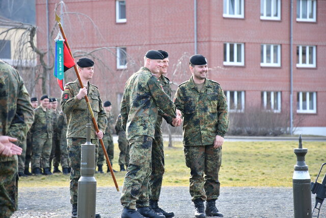 Auf dem Kasernengelände findet die feierliche Übergabe der 2. Kompanie des Panzerbataillons 393 statt. Im Vordergrund schütteln zwei Soldaten in Flecktarn-Uniformen und schwarzen Baretten die Hände. Beide lachen und blicken sich freundlich an. Im Hintergrund stehen mehrere Soldaten in Reih und Glied, ein Soldat hält eine Fahnenstange mit einem grünen und roten Wimpel. Die Szene spielt sich vor einem Kasernengebäude mit roten Ziegelmauern ab.