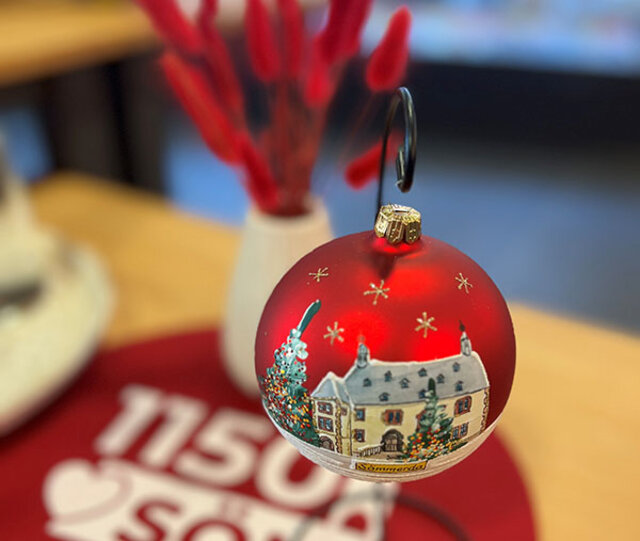 Eine rote, matt glänzende Kugel hängt an an einem kleinen Ständer. Kleine goldene Sterne rahmen das auf die Kugel kunstvoll gemalte Sömmerdaer Rathaus und eines geschmückten Weihnachtsbaums. Im Hintergrund steht ein Strauß mit rot gefärbten Gräsern.