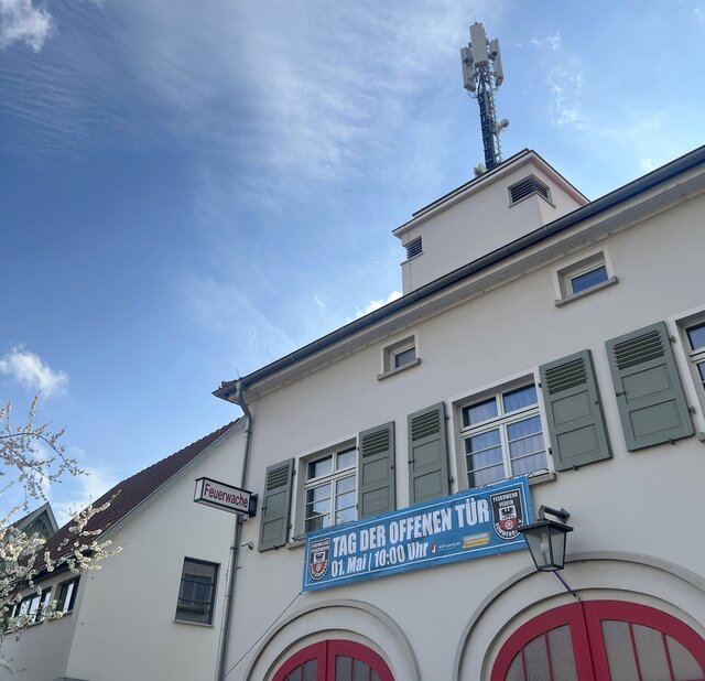 Ein Teil eines Gebäudes der Feuerwache Sömmerda mit roten Rundbogen-Garagentüren sowie Sirenenanlage auf dem Dach. Und der oberen Fensterrreihe hängt ein Banner, das auf den Tag der offen Tür am 1. Mai hinweist. 