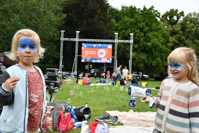 KJP-Familiensommerkino im Stadtpark Sömmerda Zwei Kinder mit geschminkten Gesichtern spielen mit Seifenblasen auf einer Wiese im Stadtpark Sömmerda, im Hintergrund ist die Bühne des KJP-Familiensommerkinos zu sehen.