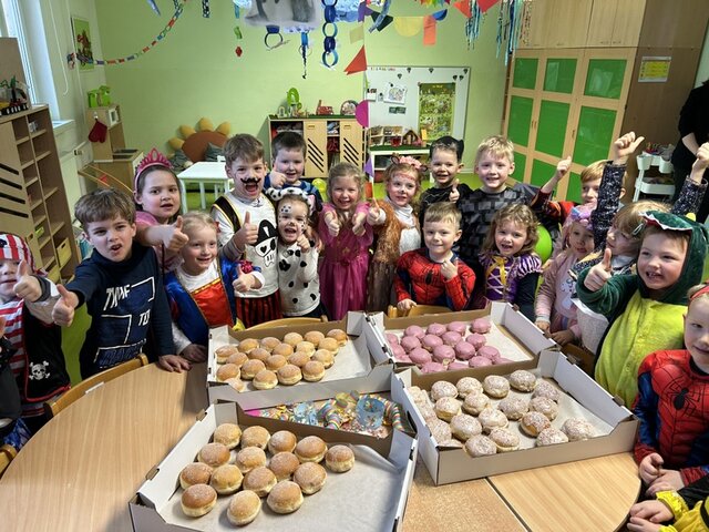 Auf einem Tisch in einem Kita-Gruppenraum stehen vier flache Kartons mit Pfannkuchen. Diese sind mit Zucker umhüllt oder haben eine weiße Glasur mit bunten Streuseln oder eine rosa Glasur. Um den Tisch herum stehen Kinder in Faschingskostümen, strecken den Daumen in die Höhe in freuen sich über die Pfannkuchen.