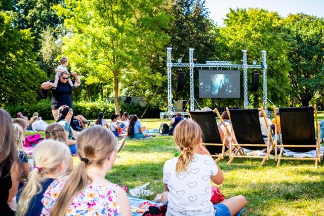 Familien sitzen bei Sonnenschein auf Decken im Stadtpark und schauen einen Film auf einer riesigen Leinwand