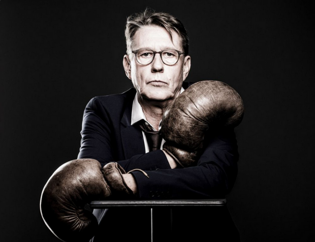 Andreas Rebers trägt einen schwarzen Anzug und Boxhandschuhe und blickt in die Kamera.