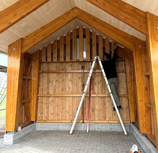 Ein offener Bau aus Holz. Ein Mann steht auf einer Leiter und schraubt bearbeitete Holzbretter an die Giebelseite des Carpot ähnlichen Baus.