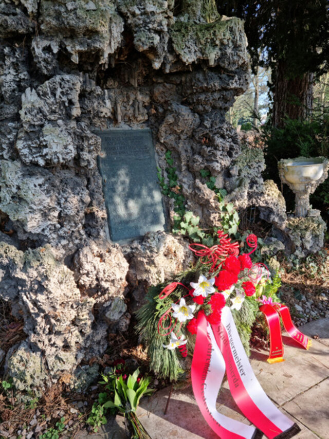 Ein Kranz mit zwei rot-weißen Bändern lehnt vor einer Gedenktafel. Diese ist an einem Gedenkstein aus Tuffsein angebracht.
