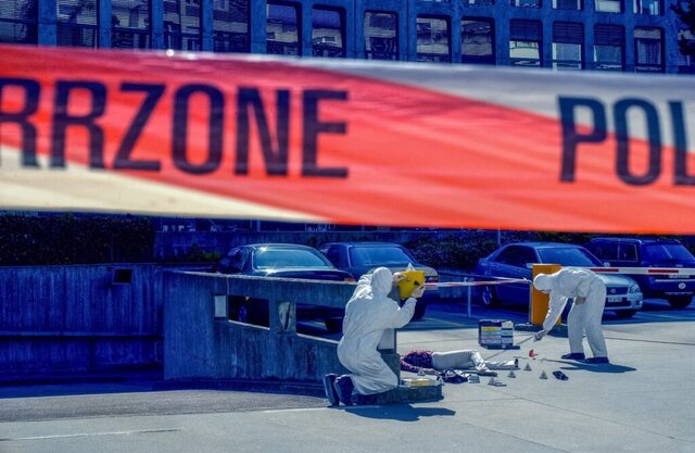 Bild zeigt 2 Menschen in weißen Schutzanzügen während der Spurensicherung vor einem großen Gebäude davor noch ein Absperrband