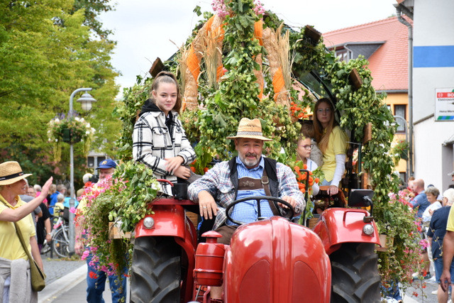 Ausschnitt vom Ernteumzug mit rotem Traktor, Fahrer und Beifahrern. Geschmückt mit Hopfen und Blumen