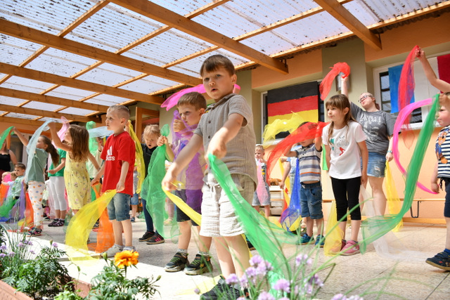 Kita-Kinder stehen auf der überdachten Terrasse des Kindergartens und schwenken farbenfrohe bunte Tücher.