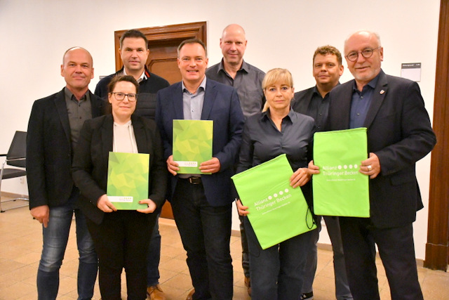 Zwei Frauen und sechs Männer stehen in zwei Reihen hintereinander im Foyer des Rathauses. Die zwei frauen und zwei Männer in der ersten Reihe halten große Umschläge bzw. Rucksäcke im Hellgrün der Allianz in den Händen, auf denen das Logo der Allianz - vier Punkte und dazu der Schriftzug Allianz Thüringer Becken - steht.