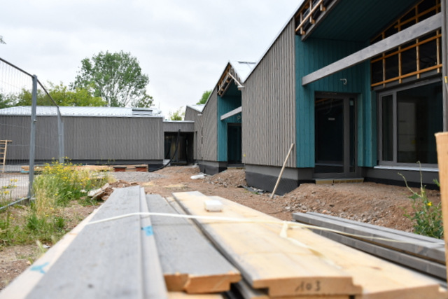 Im Vordergrund ein Stapel mit Holz-Verschalungsbrettern. Von dort aus geht der Blick zum Neubau der Kita in Patina-Grau mit petrolfarbiger Absetzung der im Bau zurückgesetzten Eingangsbereiche für die verschiedenen Altersgruppen. Der Holzneubau-Komplex bildet einen rechten Winkel. Links ist ein Bauzaun zu sehen.