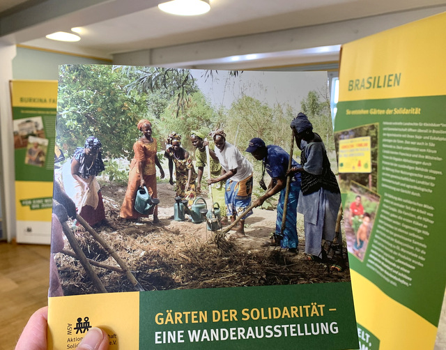 Ein Informationsblatt zur Wanderausstellung mit dem Titel "Gärten der Solidarität". Es zeigt Menschen in bunter Kleidung in einem afrikanischen Land, wie sie gemeinsam mit Hacken die trockende Erde auflockern, um etwas anzubauen.Links und rechts des Informationsblattes ist leicht verschwommen jeweils eine der Ausstellungstafeln zu sehen.
