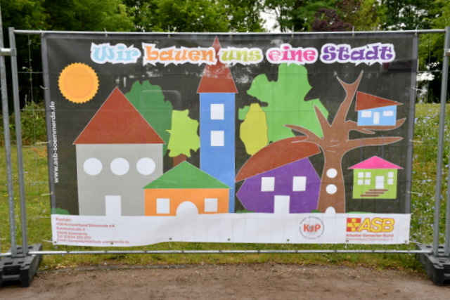 Ein Banner mit einer modellhaft dargestellten Kinderstadt in bunten Farben hängt an einem der Bauzäune.