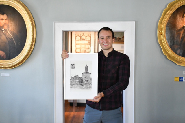 Ein junger Mann steht vor einer geöffneten Raumtür im Museum Sömmerda und präsentiert ein gerahmtes Bild mit einer Radierung, die das Erfurter Tor und einen Baum zeigt.Linkks und rechts neben dem jungen Mann hängen an der Wand historische Porträts.