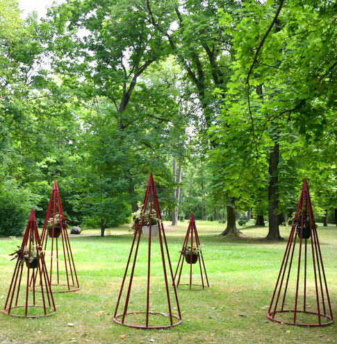 Auf einer Rasenfläche im Kronbiegelpark stehen fünf aus weinroten Metallrohren auf kreisförmiger Grundfläche zusammengefügte Tipis. In deren Spitze hängt jeweils eine bepflanzte Blumenampel. Das Foto entstand kurz nach der Installation der Tipis.