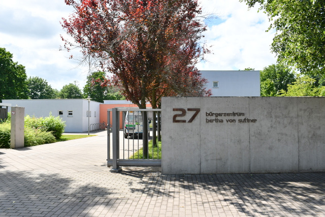 Das offene Tor führt auf das Gelände des Bürgerzentrums mit dem zweistöckigen Gebäude im Hintergrund. An der Mauer aus Sichtbeton rechts neben der Toreinfahrt ist die Hausnummer 27 angebracht sowie der Schriftzug Bürgerzentrum Berta von Suttner. Im Hof des Bürgerzentrums steht ein Kastenwagen und sind verschiedene Bäume zu sehen.