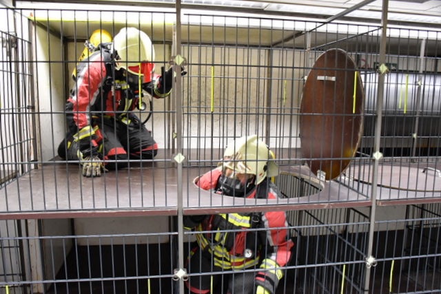 Zwei Feuerwehrleute in Einsatzkleidung und mit Atemschutzgerät befinden sich in in der Atemschutzstrecke, die einem zweietagigen Käfig, der nur auf allen Vieren absolviert werden kann, gleicht.