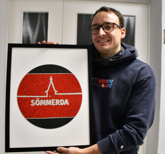 Ein junger Mann hält ein großes Bild hoch. In schwarzem Rahmen auf weißem Hintergrund zeigt es eine runde Scheibe in den farben schwarz und rot mit stilisierter Silhouette von Kirchturm und Stadtmauer. Der junge Mann trägt eine Brille und hat einen dunkelgrauen Kapuzenpullover an. 