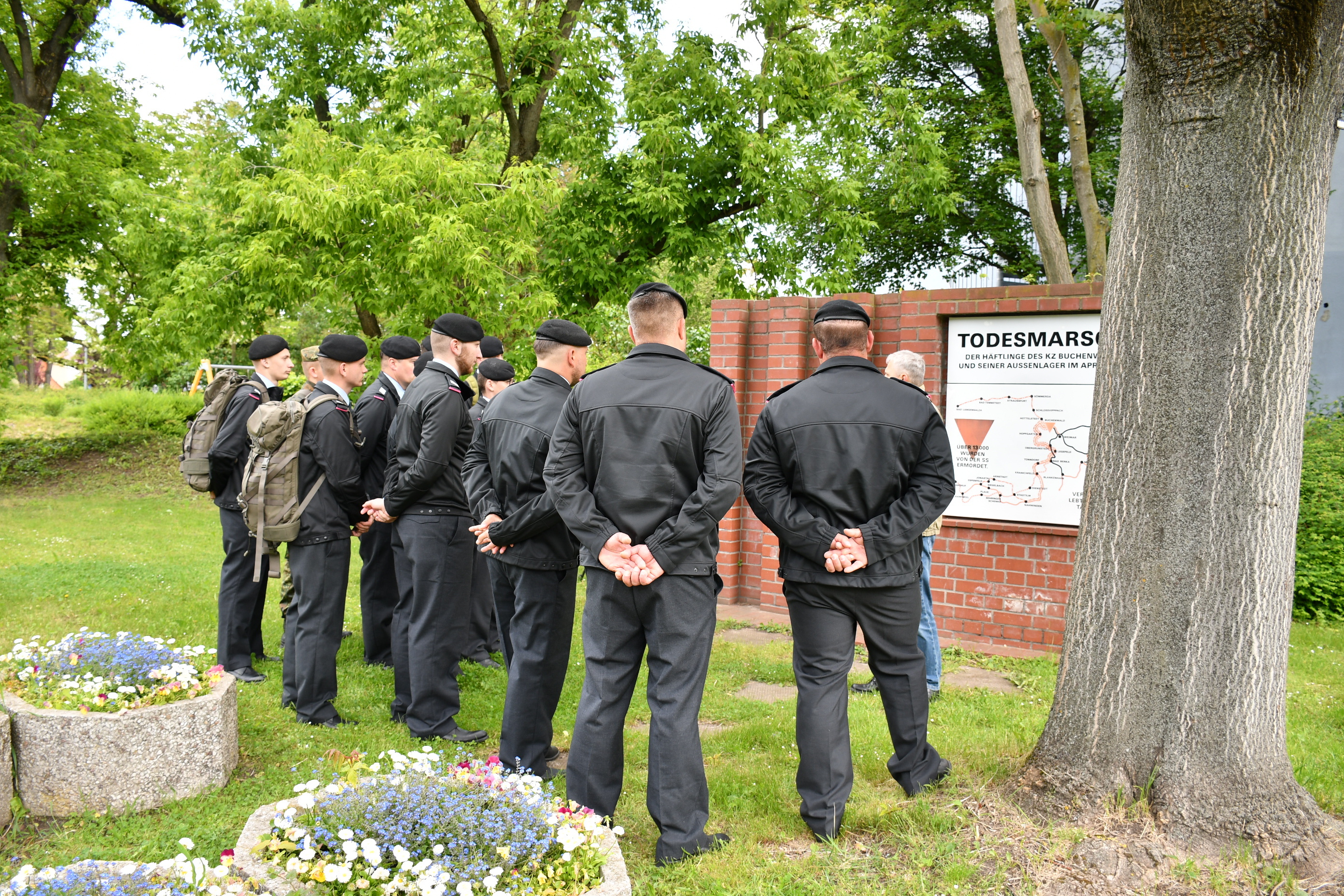Eine Gruppe von Soldaten der Bundeswehr steht in Uniformen und mit Baretten vor der Todesmarsch-Gedenkstele in Sömmerda. Sie hören aufmerksam einer Person zu, die vor der Stele spricht. Auf der Stele ist eine Karte mit der Route des Todesmarsches der Häftlinge des KZ Buchenwald im April 1945 abgebildet. Im Vordergrund blühende Blumenschalen, im Hintergrund Bäume.