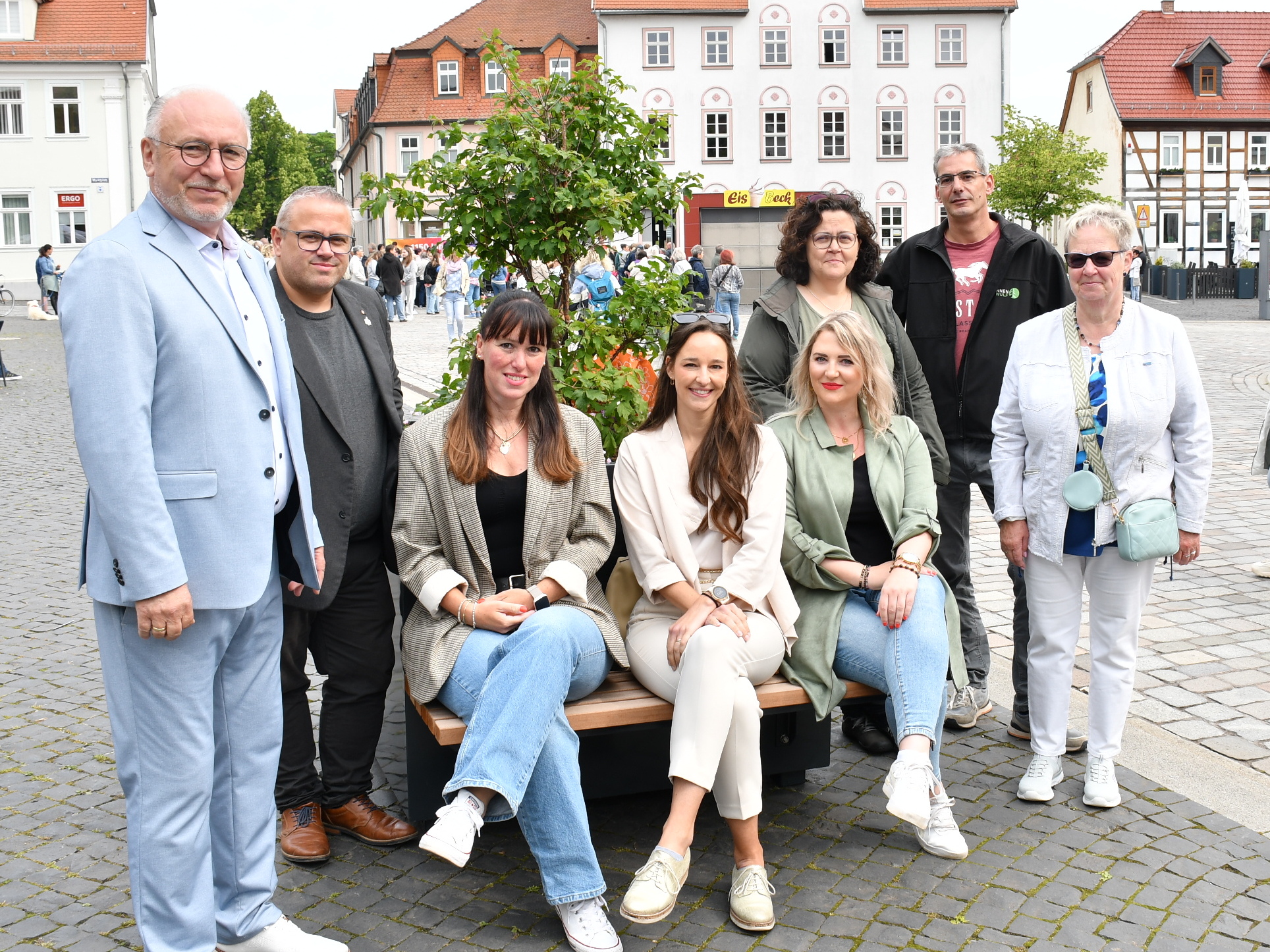 Gruppenfoto vor einem Pflanzkübel mit integrierter Sitzbank auf dem Rathausvorplatz Sömmerda. Bürgermeister Ralf Hauboldt (links im Bild) steht gemeinsam mit Projektbeteiligten und Unterstützerinnen und Unterstützern des Modellprojekts „Wanderbaumallee“. Im Hintergrund sind Marktbesucherinnen und -besucher sowie die historische Bebauung des Marktplatzes zu sehen.