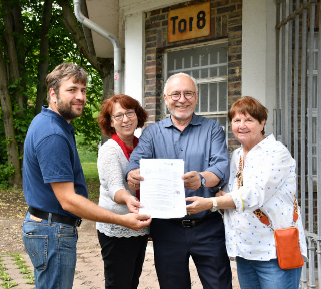 Zwei Männer und zwei Frauen stehen in einer Reihe vor einem Gebäude mit dem Schild Tor 8 und halten gemeinsam das Schreiben der Staatskanzlei mit der Fördermittelzusage in der Hand.