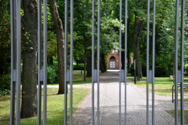 Durch die eisernen Metallzaunstreben vom geschlossenen Friedhofstor in Sömmerda ist der Hauptweg mit Blick auf die aus rotem Ziegelstein gemauerte Trauerhalle zu sehen.