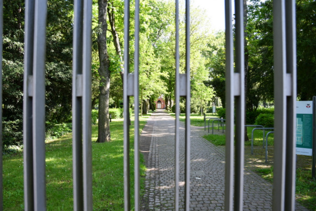 Das geschlossene Friedhofstor. Durch die Streben sieht man auf den Hauptweg des Friedhofs, an dessen Ende die Trauerhalle steht. Das Foto wurde im Sommer aufgenommen, deshalb tragen die Bäume noch Blätter.