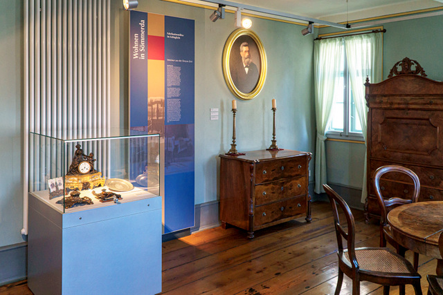 Einer der Räume der historischen Ausstellung im Museum. In dem Raum stehen eine Vitrine mit kleineren Exponaten, eine Kommode, Schrank, Tisch und Stühle. An der Wand hängen eine Erläuterungstafel sowie das Porträt eines Mannes mit Bart.