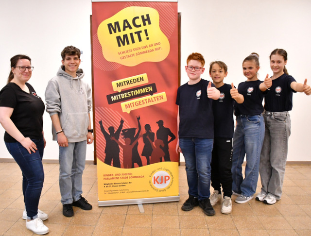 Vier neue Mitglieder des Kinder- und Jugendparlaments stehen mit erhobenem Daumen rechts vom Plakat, auf dem für eine Mitgliedschaft im KJP geworben wird. Links vom Plakat stehen der KJP-Vorsitzende sowie die Ansprechpartnerin bei der Stadtverwaltung für das KJP.