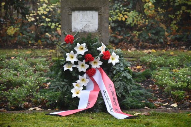 Ein großer Kranz mit Blumen in Rot und in Weiß sowie einer Schleife in Rot-Weiß liegt vor einem Mahnmal auf dem Sömmerdaer Friedhof.