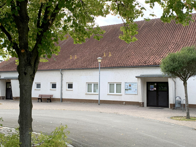 Die Sporthalle Frohndorf ist ein weißes langgestrecktes, einstöckiges Haus mit einem Spitzdach. Der Blick aufs Haus wird durch das Grün von Bäumen links und rechts eingerahmt.