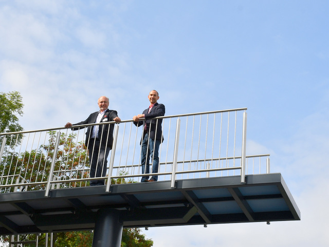 Sömmerdas Bürgermeister und Böblingens Oberbürgermeister stehen nebeneinander am Geländer auf der Plattform des Sprungturms im Stadtbad. Der Himmel ist blau mit weißen Wölkchen. Im Hintergrund leuchten grüne Blätter von Bäumen.
