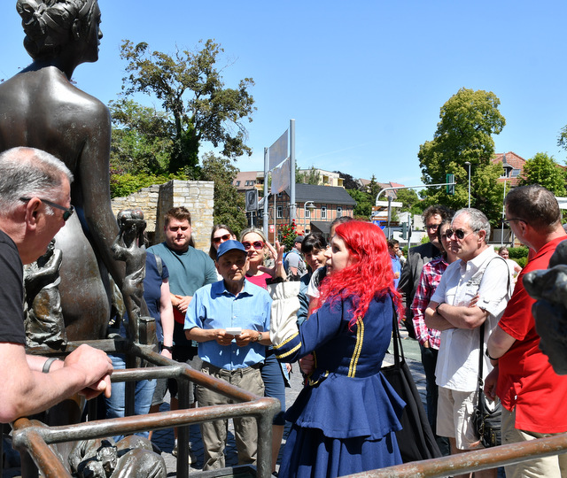 An einer Bronzeskulpturengruppe steht eine Gruppe Menschen. Die Stadtführerin erläutert ihnen das Kunstwerk.