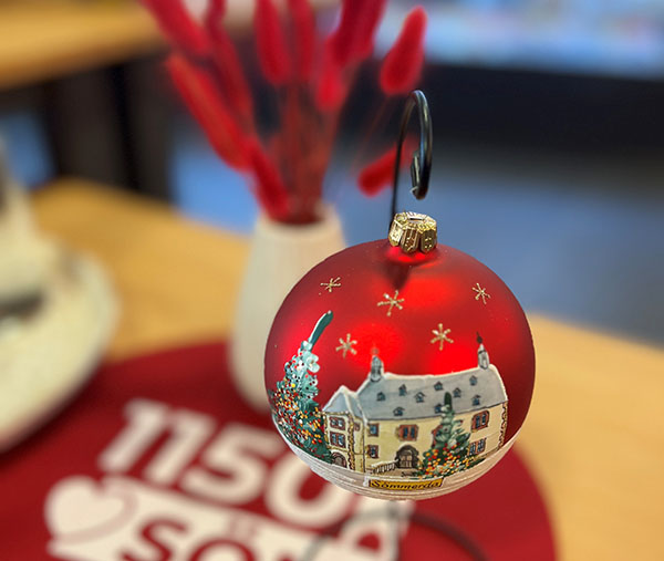 Die exklusive Weihnachtskugel mit Rathaus-Motiv und Weihnachtsbaum. Eine rote, matt glänzende Kugel hängt an an einem kleinen Ständer. Kleine goldene Sterne rahmen das auf die Kugel kunstvoll gemalte Sömmerdaer Rathaus und eines geschmückten Weihnachtsbaums. Im Hintergrund steht ein Strauß mit rot gefärbten Gräsern.