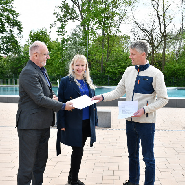 Auf der Badterrasse stehten sich Bürgermeister Hauboldt (l.) und Marco Wendelin gegenüber. Herr Wendelin hält bereits ein Blatt mit der Aufzählung gastronomischer Einrichtungen in der Hand. Der Bürgermeister übergibt ihm ein nächstes Blatt. In der Mitte zwischen beiden, leicht im Hintergrund stehend, verfolgt Laura Scherzberg das Prozedere.